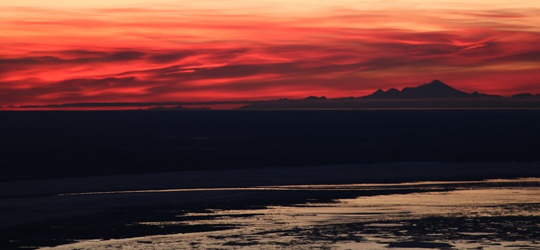 Panoramic sunset over calm water with dark shoreline and distant mountains under red and orange skies, used as a featured image for a commercial insurance blog post.