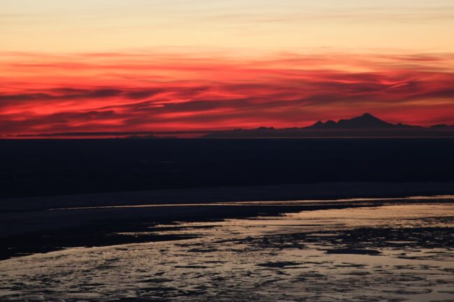 Panoramic sunset over calm water with dark shoreline and distant mountains under red and orange skies, used as a featured image for a commercial insurance blog post.